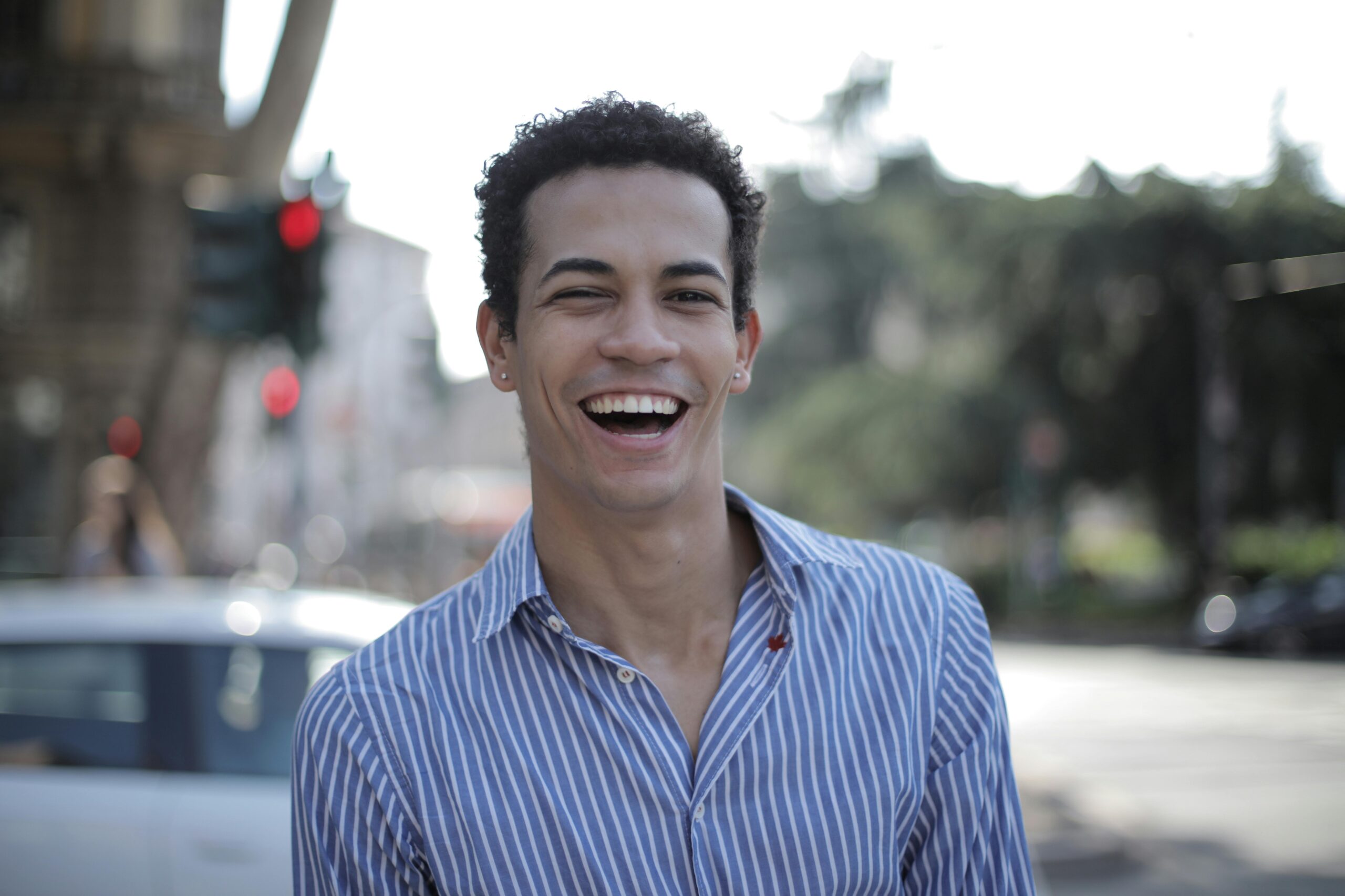 A cheerful young man in a striped shirt smiles brightly outdoors, conveying happiness and style.